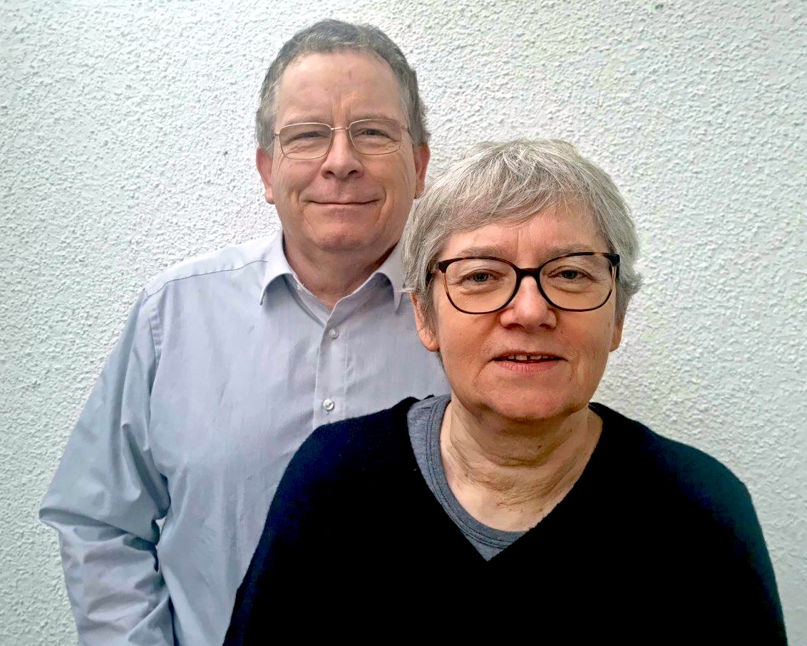 Werner und Marilies stehen vor einer weißen Wand und lachen in die Kamera. Sie tragen beide eine Brille und haben graue Haare. Werner trägt ein graues Hemd und Marilies einen dunkelblauen Pullover.